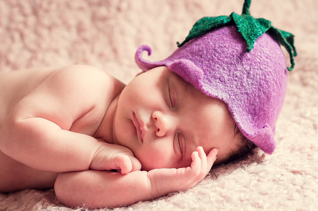 A close-up photo of a sleeping baby wearing a purple fruit hat