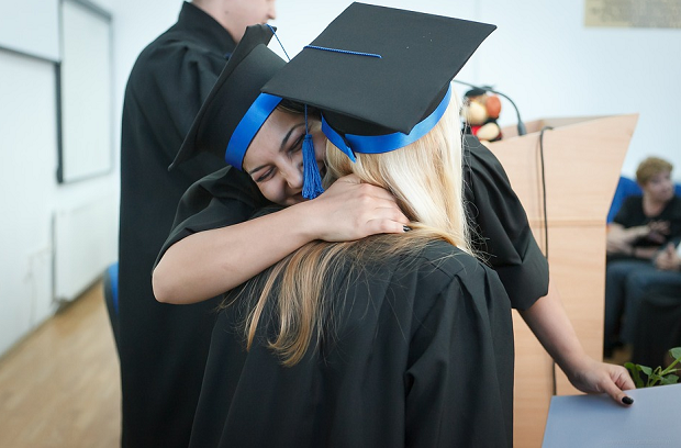 One graduate giving another a hug while someone else talks at the podium behind them