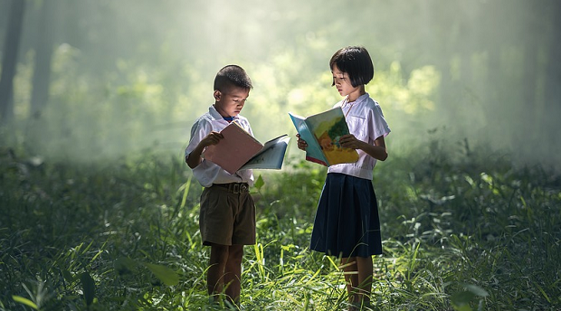 A couple of kids standing and reading books in the middle of a forest