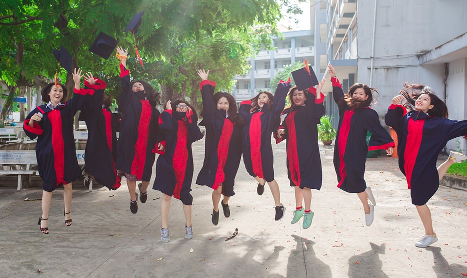 A lined up group of graduates in black and red gowns jumping and throwing their hats