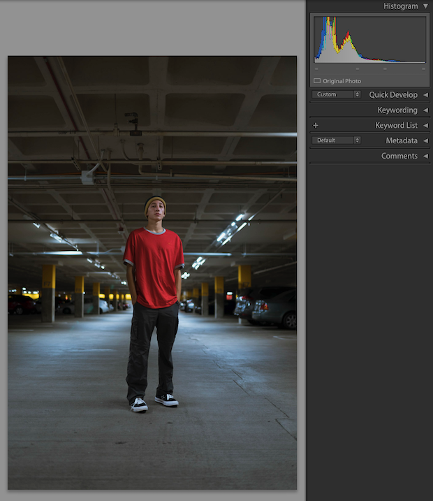 Man in jeans and a red t-shirt standing in the center of a parking garage with cars on either side of him.