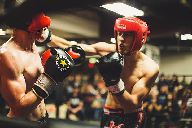 Two boxers in the middle of a round. One boxer faces the camera and punches with one arm completely outstretched. The other boxer's back is to the camera, though slightly turned so his chest is visible but not his face.