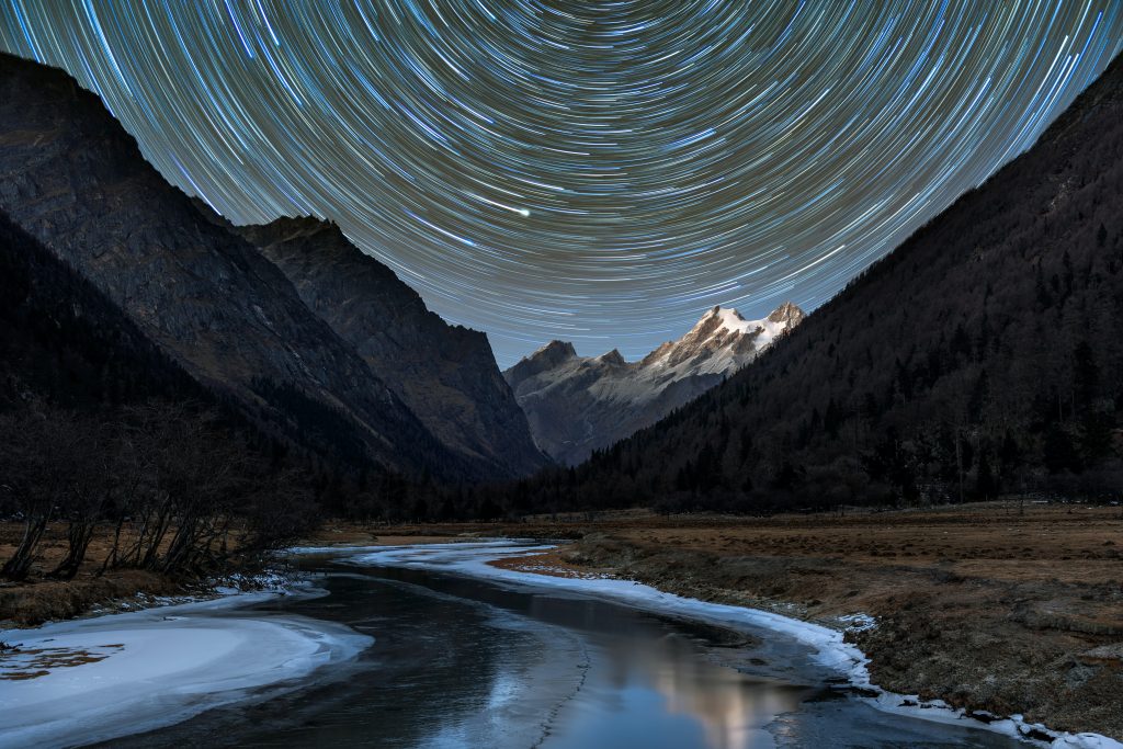 A frozen river flowing alongside snowy the mountains with circular star trails at the background