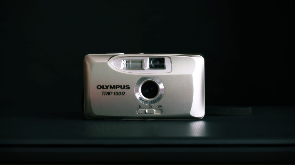 A silver-colored Olympus Trip 100R camera placed at the center of a black tabletop in front of a black background 