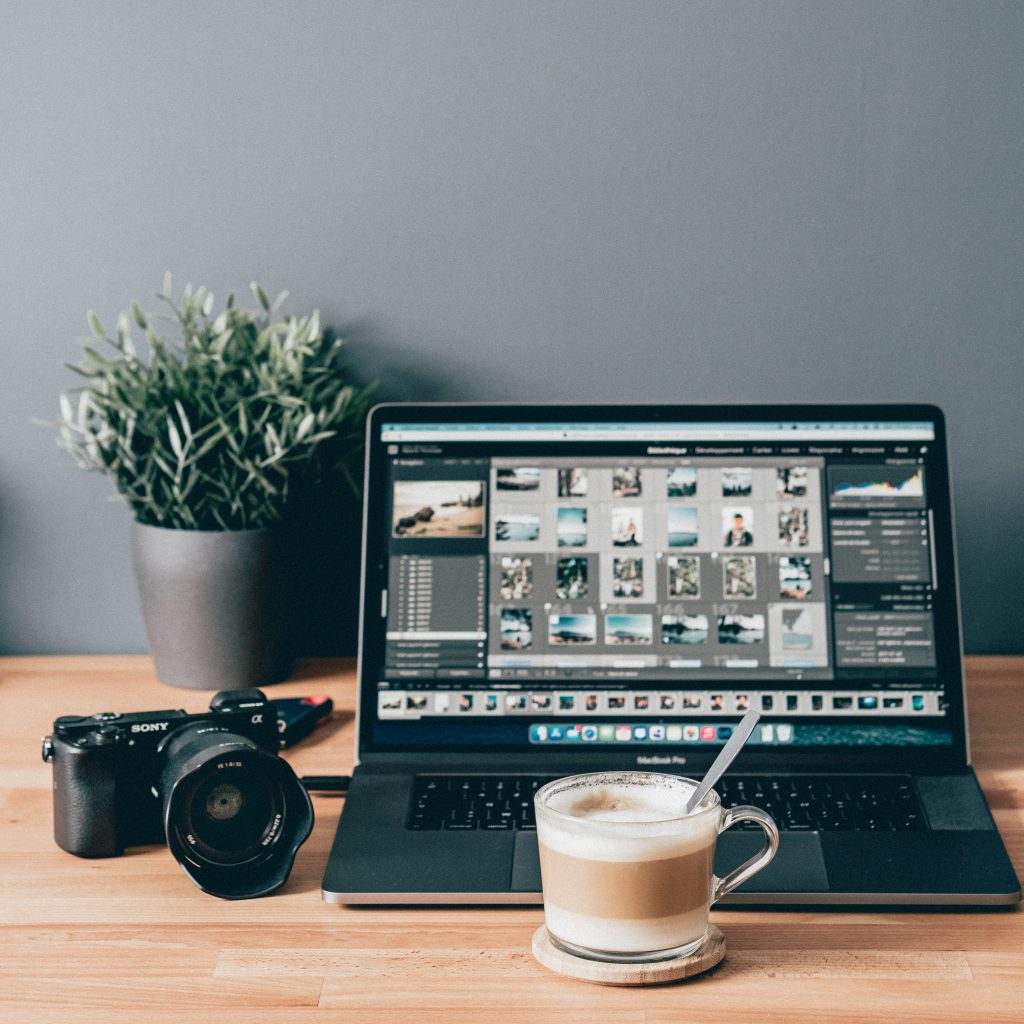 A camera, a coffee mug, a laptop displaying Lightroom catalog placed on top of a wooden desk